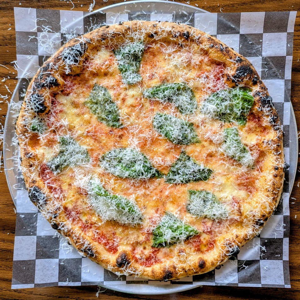 Wood-fired cheese pizza topped with basil pesto and grated cheese on black-and-white checkered paper.