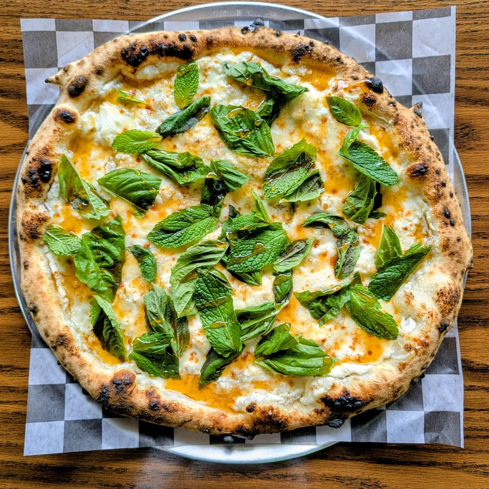Wood-fired pizza topped with melted cheese, orange sauce, and fresh mint leaves on checkered paper.