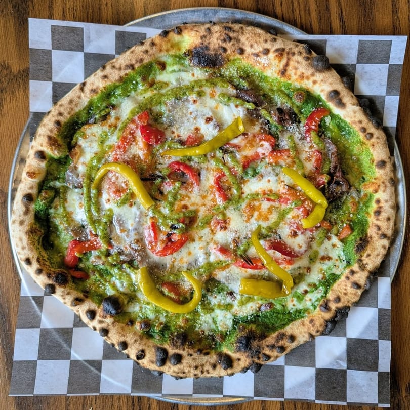 Pop-Start Pizza's pesto pizza with red peppers and yellow banana peppers on a checkered paper.